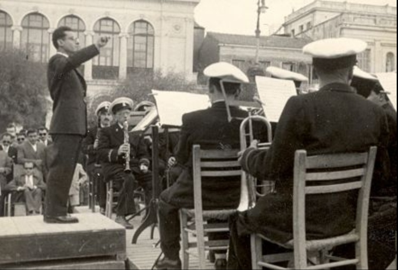 30. Ο Θεόφιλος Κάβουρας (1930-1974). Ο μαέστρος διευθύνει την μπάντα τού Δήμου Πατρέων στην Πλατεία Γεωργίου. Στο φόντο διακρίνεται (αριστερά) το Δημοτικό Θέατρο και το κτίριο της Εθνικής Τράπεζας (δεξιά).jpg