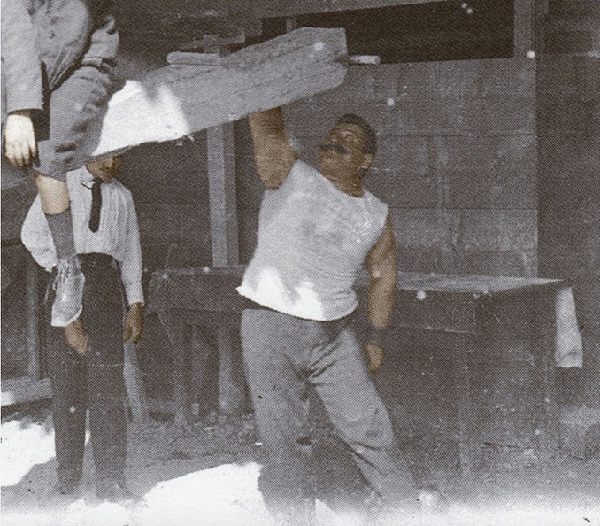 5. Ο Δημήτρης Τόφαλος σηκώνει με το ένα χέρι ένα ξύλινο δοκάρι που κάθονται πάνω του δύο μικρά παιδιά. (επιχρωματισμένη λεπτομέρεια από φωτογραφία πριν από το 1906).jpg